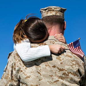 A soldier in uniform hugged by another holding an American flag against a blue sky- Moss Bollinger LLP