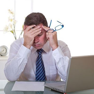 A stressed person at a desk with a laptop, holding their head and glasses- Moss Bollinger LLP