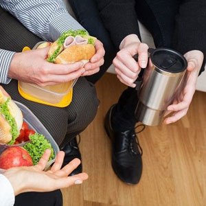 Two people in formal attire sitting with a sandwich, plastic container, mug, plate with food including sandwich, apple, and broccoli- Moss Bollinger LLP