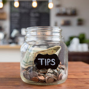 A “TIPS” jar filled with coins and bills on a wooden surface in a cafe or eatery- Moss Bollinger LLP