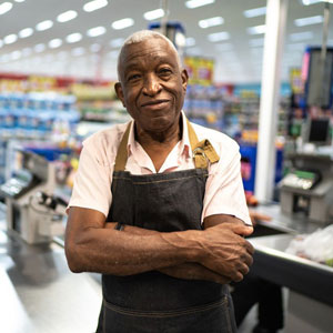 Person in white shirt and dark apron, arms crossed at store counter with well-lit, stocked shelves- Moss Bollinger LLP